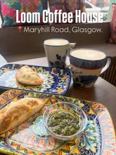 A high-angle shot of a table at Loom Coffee House featuring two empanadas, a bowl of chimichurri, and two coffees in patterned mugs. The image captures a casual, cozy cafe setting with natural window lighting.