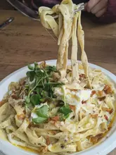 A close-up, action-oriented shot of a fork lifting a portion of truffle linguine from a disposable plate, topped with crispy onions and pea shoots.