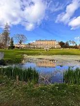 A scenic, wide-angle exterior shot of a large, historic manor house situated behind a pond, captured under a bright blue sky.