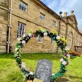 A photograph of an Easter-themed floral archway installation positioned on the grounds in front of a historic stone building, likely Harewood House, intended to promote seasonal family-friendly activities.