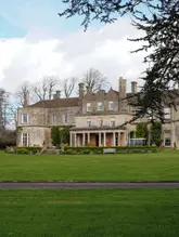 A wide, exterior shot of a historic country house hotel, Lucknam Park, set against a green lawn and overcast sky.
