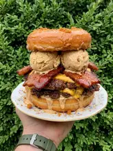 A close-up, vertical shot of an experimental 'Frankenfood' creation featuring a double smash burger with bacon, cheese, and scoops of Biscoff ice cream sandwiched between two glazed donuts. The image is held by a hand against a green hedge background.