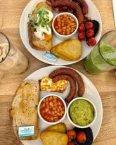 An overhead flat-lay shot of a brunch spread at a cafe, featuring two plates of breakfast items including eggs, sausages, bacon, halloumi, beans, hash browns, and vegetables, accompanied by an iced coffee and a green smoothie.