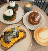A high-quality, overhead flat-lay photograph of a brunch spread featuring poached eggs on toast, scrambled eggs with mushrooms, a cinnamon roll, and two coffees, set on a wooden table.