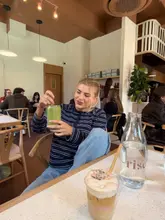 A lifestyle-focused shot capturing a customer enjoying a matcha drink in a bright, modern cafe setting. The image conveys a relaxed, social brunch atmosphere, highlighting the venue's interior and the coffee shop experience.