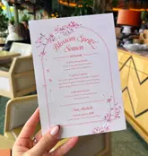 A first-person perspective shot of a printed 'Blossom Spritz Season' menu held in a hand, with a blurred, upscale restaurant interior in the background.