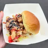 A close-up, handheld shot of a chicken burger served in a cardboard tray, captured at a food festival event. The image focuses on the textures of the fried chicken, sesame seeds, and sauce, presenting the food in a casual, authentic manner typical of street food reviews.
