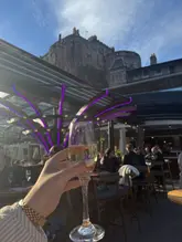 A first-person perspective shot holding a glass of sparkling wine against the backdrop of Edinburgh Castle from a rooftop terrace. The image captures the atmosphere of the venue, highlighting the view and the outdoor setting.