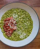 A high-quality, overhead shot of a homemade lemon and spinach risotto topped with crispy prosciutto and parmesan shavings. The image is well-lit with natural light, showcasing the vibrant green colour of the risotto and the texture of the crispy meat.