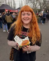 A casual, candid-style photo of a creator holding a burger at an outdoor food festival event. The image captures the atmosphere of the venue but lacks the technical precision required for high-end food marketing.