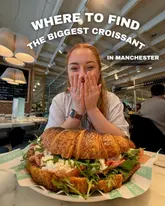 A bright, engaging promotional image featuring a creator holding a giant croissant sandwich in a restaurant setting. The composition is centered and clear, effectively highlighting the 'biggest croissant' hook mentioned in the text overlay. The lighting is even and the food appears fresh and appealing, making it a strong piece of social content for a hospitality venue.