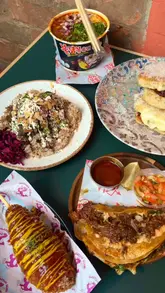 A montage of various food items from different vendors at a food hall, featuring close-up shots of birria ramen, tacos, a focaccia sandwich, a lamb rice box, a Korean hot dog, and taco pancakes.