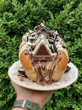 A close-up, eye-level shot of a novelty 'dessert hot dog'—a donut bun filled with chocolate sauce, topped with a Toblerone piece and crushed Oreo cookies—held on a vintage saucer against a dense green hedge background.