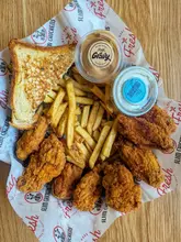 A top-down, well-lit photograph of a fried chicken meal from Slim Chickens, featuring chicken tenders or wings, fries, a slice of Texas toast, and two dipping sauces in a branded basket.