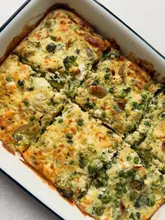 A top-down, well-lit photograph of a freshly baked green vegetable frittata, cut into squares within a white enamel baking dish. The dish contains broccoli, peas, leeks, potatoes, and feta cheese, showing a golden-brown, crispy top.