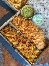 A high-quality, top-down photograph of a classic fish and chips meal served in a branded takeaway box, accompanied by mushy peas and tartare sauce.