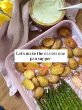 A bright, top-down shot of a home-cooked salmon and potato traybake, accompanied by roasted asparagus and a creamy dipping sauce. The food is presented in a pink marble-effect tray on a wooden surface, styled with a soft cloth and fresh flowers, creating an inviting and clean aesthetic.