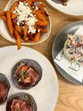 A top-down, overhead shot of a brunch spread featuring tuna tacos on blue corn tortillas, an iceberg lettuce wedge salad, and sweet potato fries topped with feta, set on a wooden table.