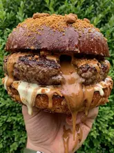 A close-up, high-contrast shot of a 'Biscoff' stuffed burger being held in a hand against a natural green background. The image focuses on the 'lava' effect of the sauce oozing from the center of the patty, emphasizing the messy, indulgent nature of the food.