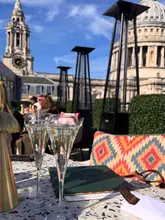A bright, sunny, eye-level shot taken from a table at a rooftop venue, featuring two glasses of sparkling wine in the foreground with a menu, set against the iconic backdrop of St. Paul's Cathedral.