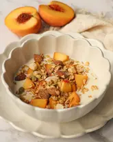 A high-quality, bright, and inviting food photography shot featuring a bowl of granola with coconut yogurt and fresh peach slices. The composition is clean and symmetrical, with soft, natural lighting that highlights the textures of the ingredients.