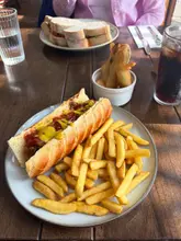 A casual, brightly lit shot of a hot dog, fries, and a side of fried pickles served on a wooden table, likely at a restaurant with a view.