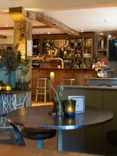 A high-quality, atmospheric interior shot of a pub, capturing the warm, inviting ambiance of the space. The composition highlights the bar area and a cozy foreground table setting, effectively conveying the venue's character.
