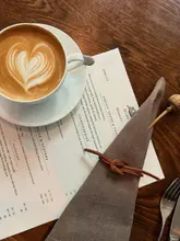 A high-quality, overhead flat-lay shot of a breakfast setting at a restaurant, featuring a latte with intricate heart-shaped latte art, a printed menu, and a neatly folded napkin on a wooden table.