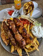 A bright, high-angle shot of a Greek meal featuring a plate of souvlaki skewers, fries, pita, tzatziki, and grilled feta, with a gyros wrap visible in the background on a wooden table.