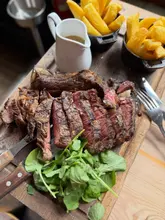 A high-quality, appetizing close-up shot of a sliced steak served on a wooden board with a side of chips and a gravy boat, captured in a rustic, inviting setting.