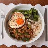 A top-down, well-lit photograph of a homemade Pad Kra Pao dish served in a white bowl on a placemat, featuring minced pork, runner beans, white rice, tenderstem broccoli, and a fried egg topped with chilli flakes.