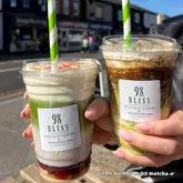 A high-quality, bright, and inviting shot of two specialty iced drinks held in front of a cafe, showcasing the layered textures and toppings of a baklava iced latte and a strawberry cheesecake iced matcha.