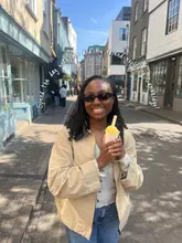 A bright, sunny 'day in the life' style photo featuring a creator holding a cup of gelato while exploring Cambridge. The image captures a casual, friendly vibe suitable for social media promotion of a day out.