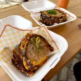 A candid, high-quality shot of two toasted sandwiches served in takeaway containers on a wooden table. The image captures the texture of the toasted bread and the melted cheese filling effectively, conveying the appeal of a casual, high-quality street food experience.