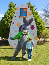 A bright, outdoor photograph of a young child posing with an Easter Bunny mascot in front of a large, polka-dot egg prop at a family-friendly park.