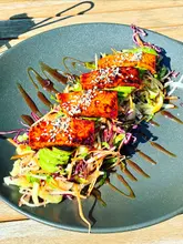 A top-down, overhead shot of a vegan tofu salad served on a dark plate, placed on a wooden outdoor table. The dish features grilled tofu, avocado, and a slaw mix, garnished with sesame seeds and a balsamic glaze.