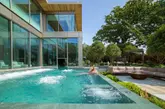 A high-quality, professional lifestyle photograph showcasing the outdoor spa facilities at Sopwell House. The image features a couple relaxing in a heated pool, with a modern glass-fronted building and lush greenery in the background, effectively communicating a premium, relaxing, and aspirational atmosphere.