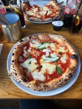 A top-down, slightly angled shot of a freshly baked Neapolitan-style pizza on a wooden table, with a second pizza and drinks visible in the background, capturing a casual dining experience.