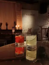 A moody, atmospheric shot of two cocktails placed on a reflective table in a dimly lit, underground speakeasy setting. The background features a brick wall with the venue name 'VICEROY' subtly illuminated, reinforcing the exclusive, hidden nature of the bar.