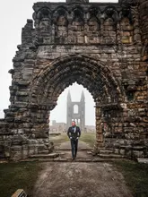 A high-quality, atmospheric photograph of a man walking through the historic ruins of St Andrews Cathedral in Scotland. The composition uses the stone archway to frame the subject, creating a sense of scale and historical depth.