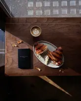 A high-quality, overhead flat-lay photograph featuring a flaky croissant, a latte with latte art, and a branded notebook on a wooden table, capturing a cozy, aesthetic bakery experience.