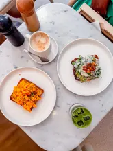 A high-quality, overhead flat-lay shot of a breakfast spread at Claridge's Artspace Cafe, featuring scrambled eggs on toast, an avocado-based dish, a coffee, and a matcha latte on a clean marble table.