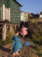 A high-quality, evocative lifestyle photograph capturing two children walking along a sandy path towards a green beach hut during golden hour. The image effectively conveys a sense of family-friendly coastal exploration and holiday nostalgia.