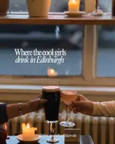 A moody, atmospheric shot of two hands toasting with a dark stout and a cocktail in a dimly lit bar, featuring a candle in the foreground.