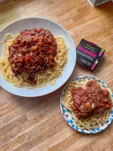 A top-down, overhead shot of two plates of spaghetti bolognese served on a wooden table, with a packet of JD Seasonings Bolognese mix placed next to the larger plate.