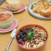 A bright, well-lit overhead shot of a cafe table featuring an acai bowl, a cheese toastie, and two hot drinks. The image captures a casual brunch setting with a focus on the fresh, colourful presentation of the food.