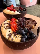 A high-quality, close-up shot of two smoothie/açai bowls served in coconut shells, featuring fresh strawberries, blueberries, bananas, and cacao nibs. The lighting is soft and natural, highlighting the textures of the fruit and the chocolate spread.