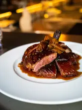 A high-quality, professional food photograph showcasing a beautifully glazed roast duck dish. The lighting accentuates the texture and gloss of the sauce, while the shallow depth of field keeps the focus entirely on the meat. The composition is clean and appetizing, effectively conveying the quality of the food at the venue.