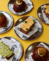 A bright, high-quality flat lay photograph showcasing a variety of Hong Kong-style baked goods from Cantoast Bakery, including honey toasts, salt breads, and bolo buns, arranged on branded paper on a vibrant yellow background.