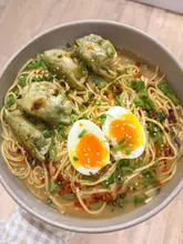 A high-quality, top-down food photography shot of a homemade noodle soup bowl featuring soft-boiled eggs, green dumplings, and scallions, presented on a clean wooden surface.